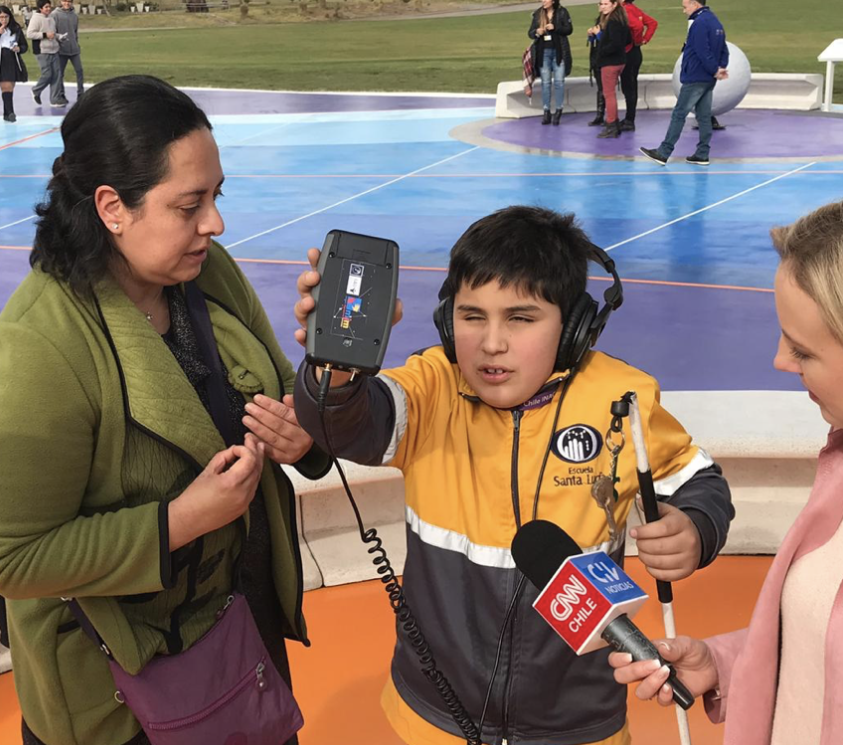 A  boy with short black hair and a yellow and black coat wearing headphones is holding a LightSound device toward the sun in one hand and his white cane in the other. He is being interviewed by a woman from CNN about his experience. Also pictured is another woman with shoulder length black hair and a green sweater. She is also asking the boy questions about his experience.