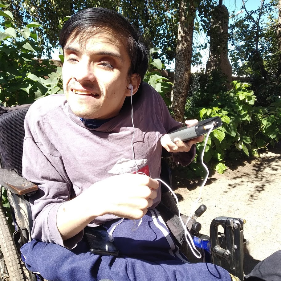 A man sitting outdoors in a motorized wheelchair holding a LightSound device up toward the Sun. He is wearing blue pants, a purple t-shirt, and listening to the device with headphones while smiling.