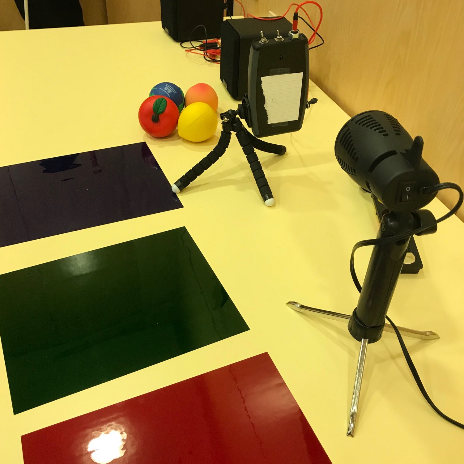 Plastic color filters (red, green, and blue) sit on a table with a LightSound device on a table top bendable tripod and a light source all ready for a demo of how the sound changes with color changes.