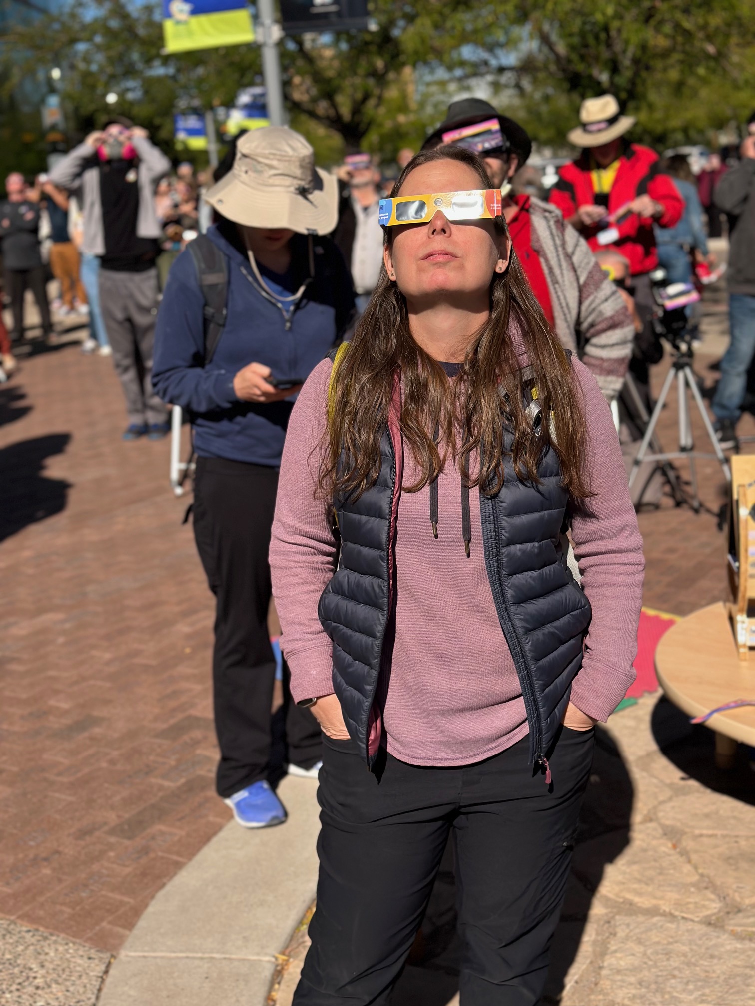 A woman standing with eclipse glases on. She is wearing black pants, a purple long sleeve shirt with a blue puffer vest. She has long brown hair and her hand are in her pockets. She is looking up at the Sun during an event of the October 14, 2023 Annular Eclipse. There are many people in the background.