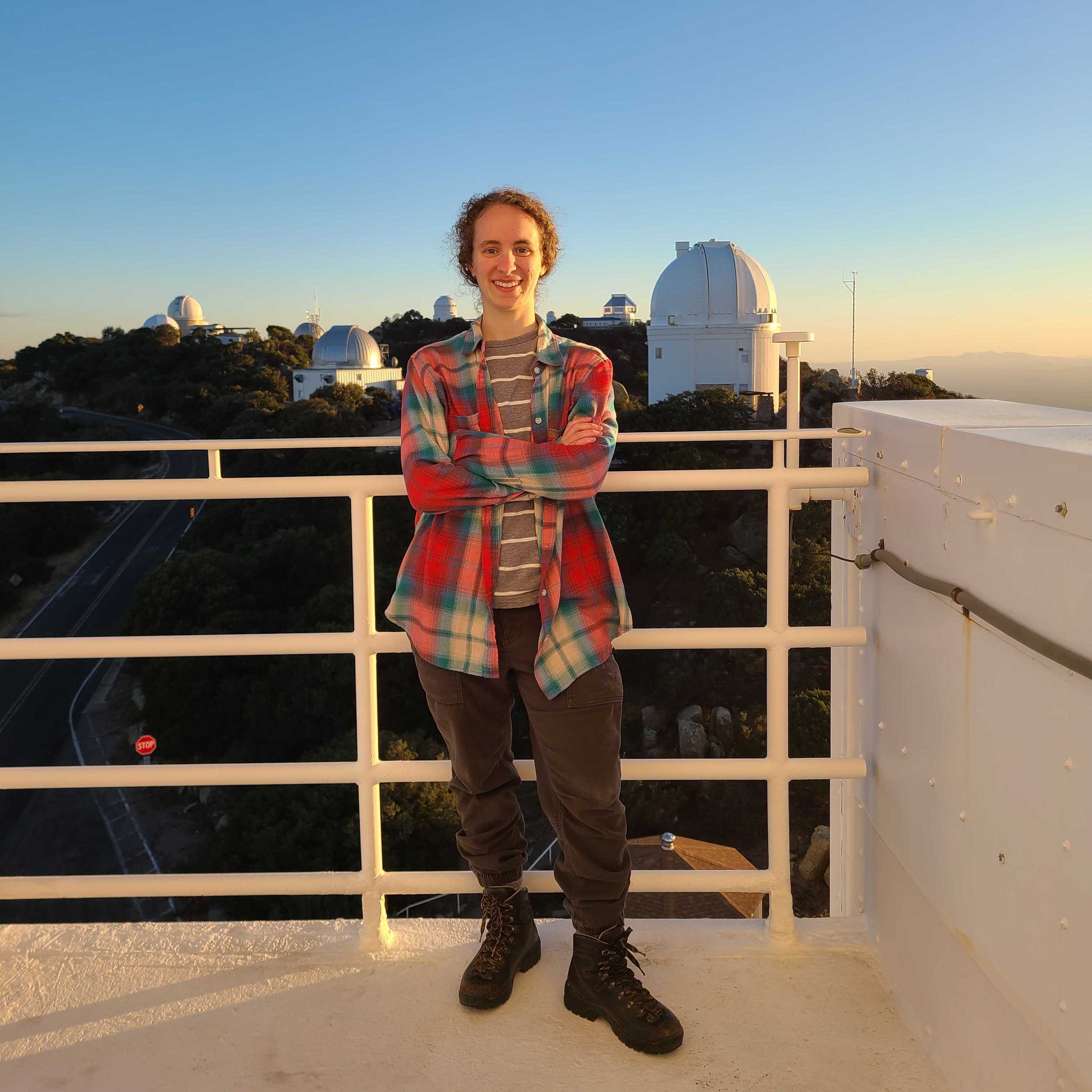 A womne with curly, dirty blond hair is standing on a balcony with a telescope dome in the background. She has dark pants and a plaid, long sleeved shirt, standing with her arms crossed and a smile on her face.
