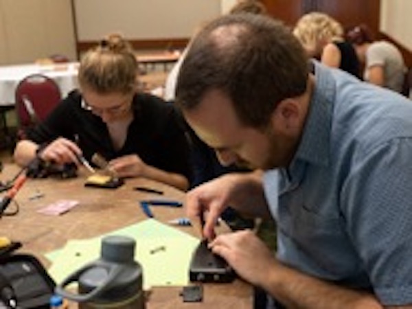 A man with a beard and a blue short sleeve shirt is assembling a LightSound case at a table with a woman who is soldering her LightSound device.