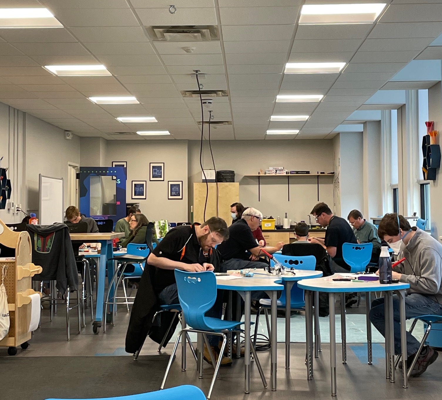 A room full of round white table and blue chairs with around ten people soldering during a soldering workshop.