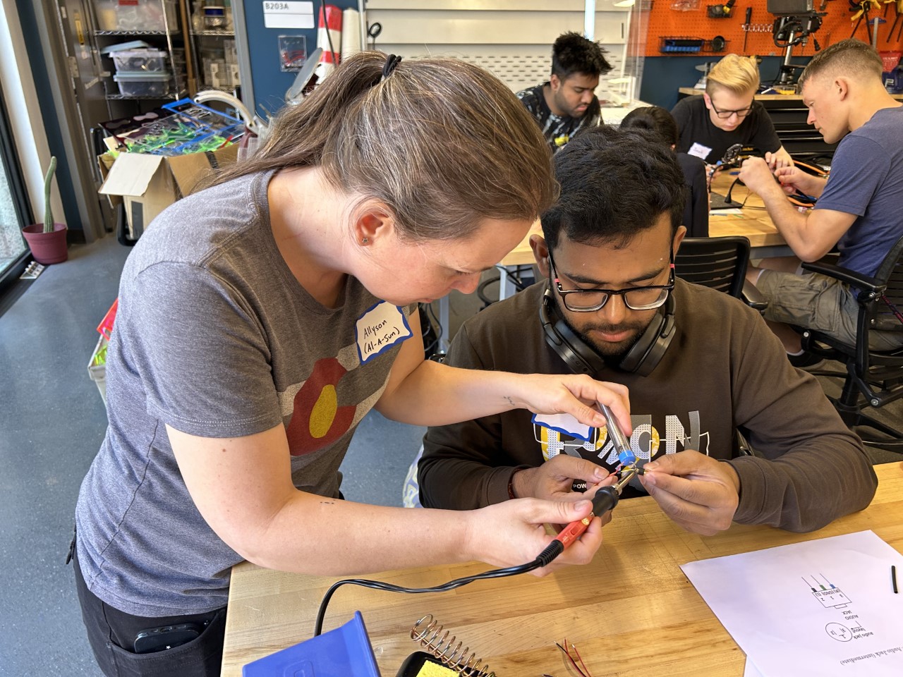 A woman in a gray Colorado flag t-shirt with a pony tail is helping a male student learn to solder. Behind them is a table of three more students soldering during a LightSound workshop.