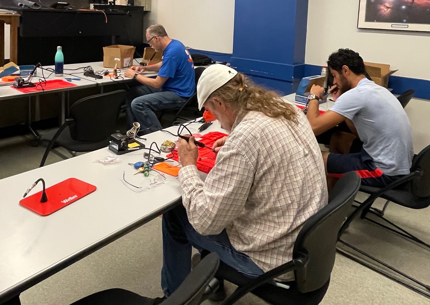 Three men shown from behind working at soldering stations to build LightSound devices at a UT Austin LightSound workshop.