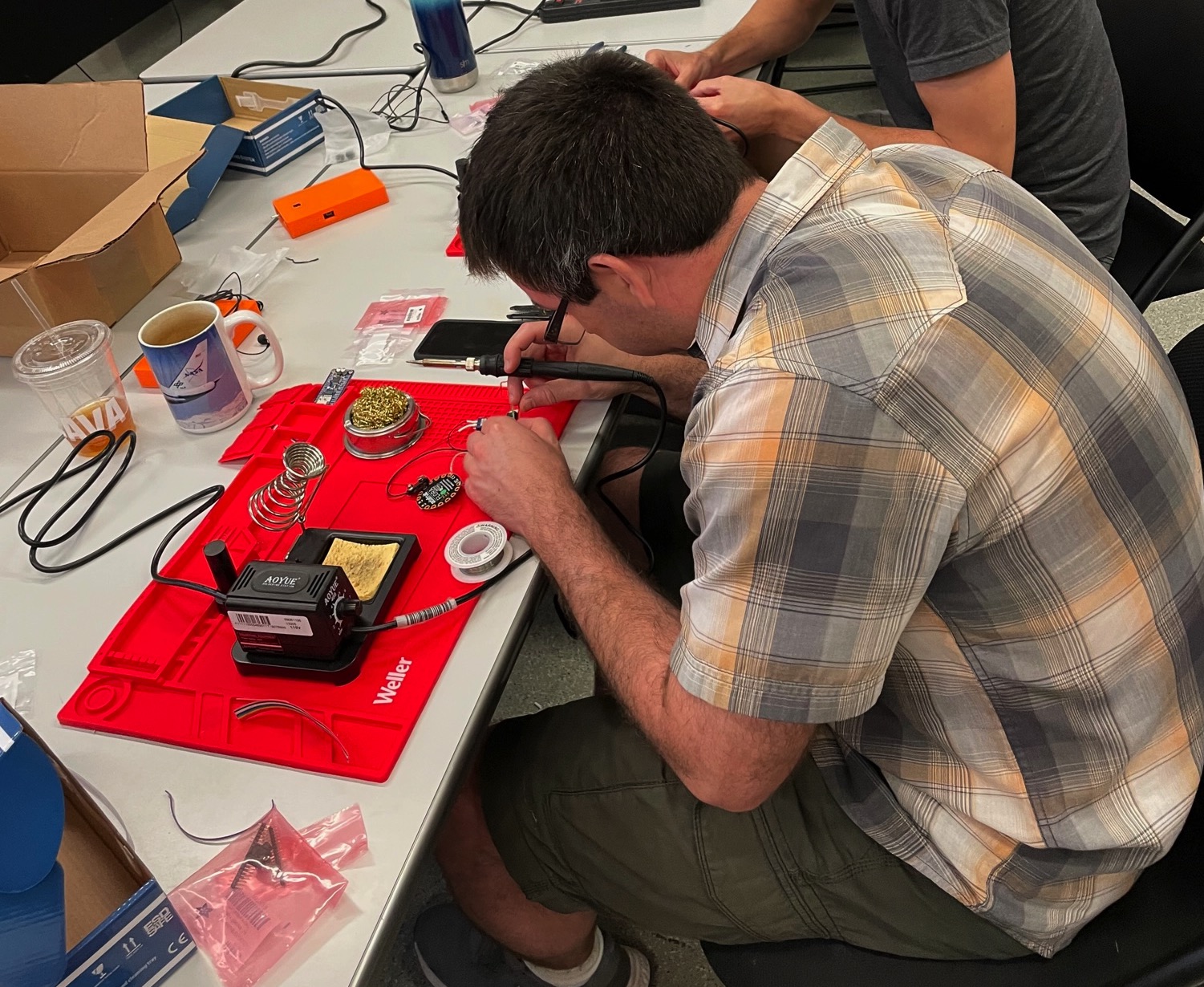 The side profile of a man in green shorts and a plaid short sleeve top wearing glasses is working at a soldering station soldering a component of the LightSound device. The table around him is scattered with supplies and a coffee mug.