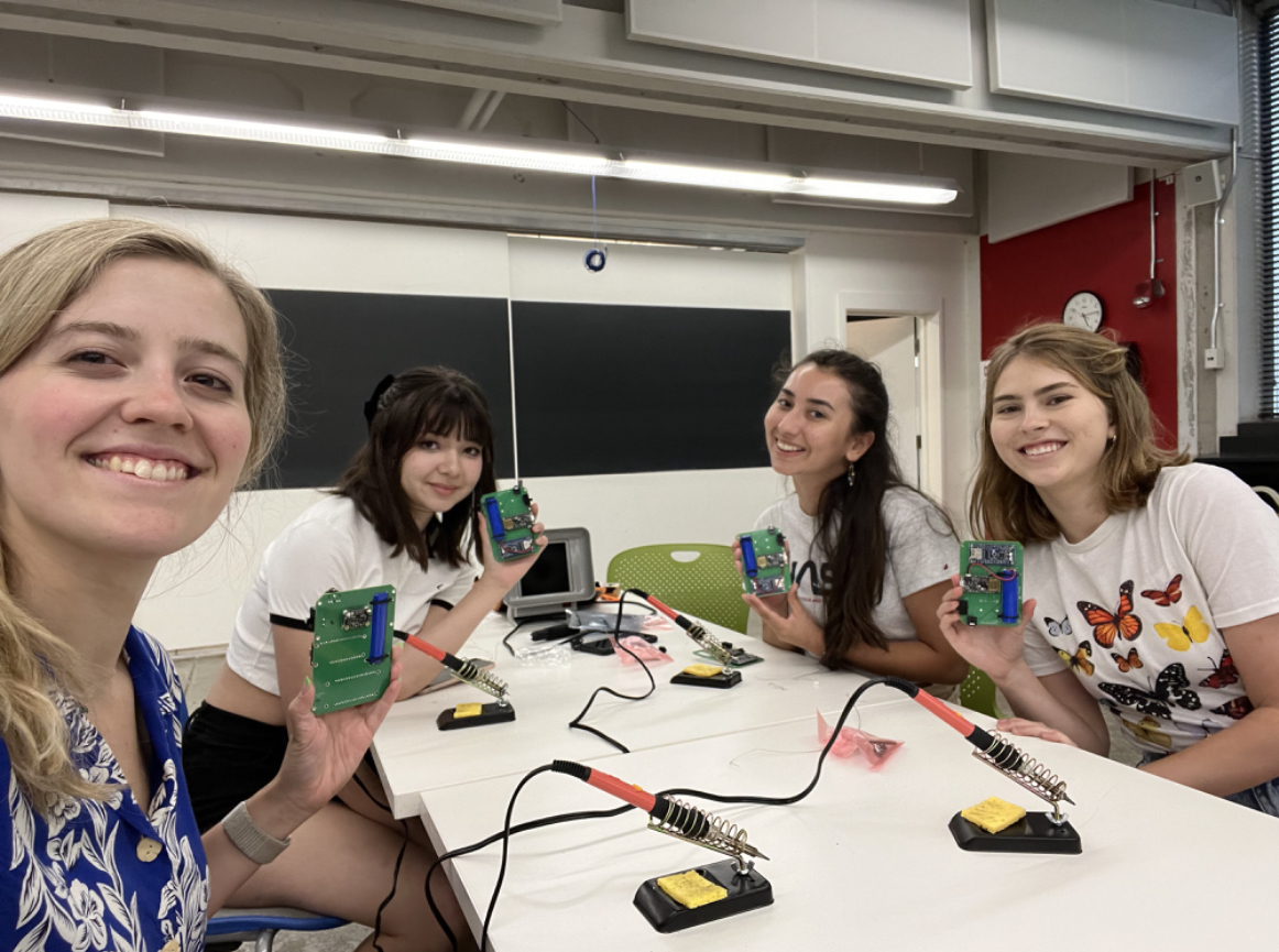 Four female students sitting around a table with soldering irons hold up their completed LightSound printed circuit boards while smiling and taking a selfie. 
