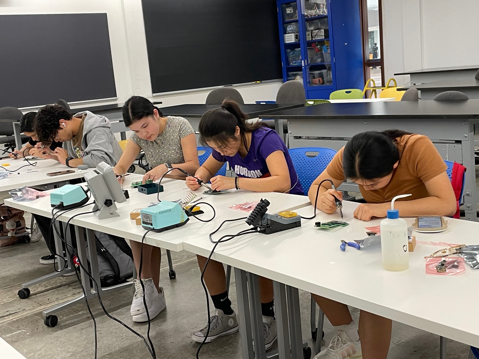A long white table with five students sitting while working at soldering stations soldering LightSound components at a Harvard University LightSound workshop.