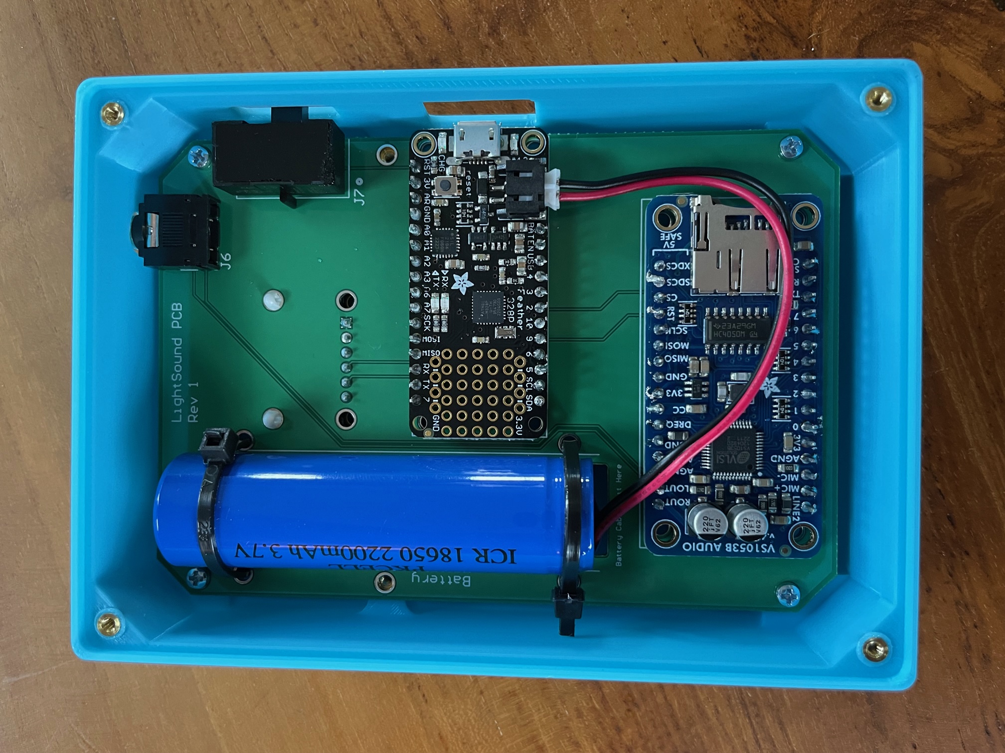 A blue LightSound case with the lid off showing the printed circuit board inside. The audio jack on the top right, on/off switch on the side right, the rectangular Feather microcontroller board in the middle, the midi board on the bottom, and a Li-ion rechargeable battery on the left of the circuit board.
